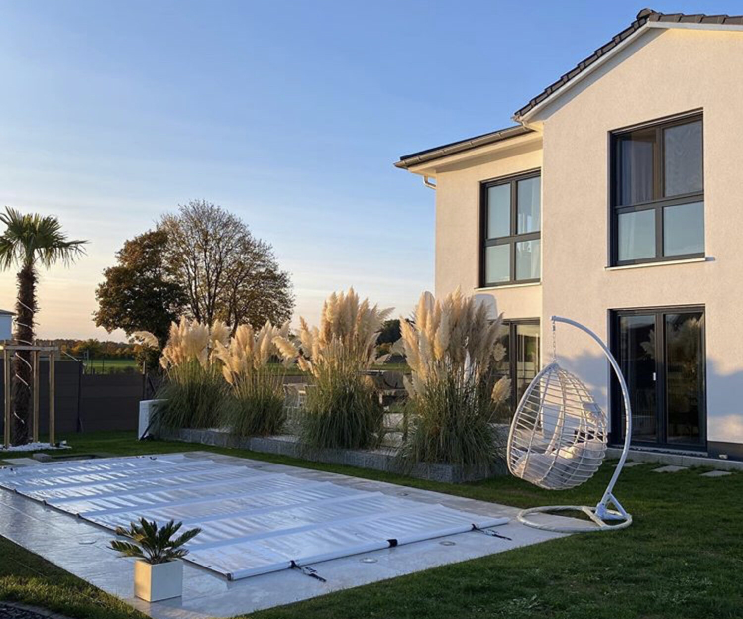 Modernes weißes Haus mit großen Fenstern, einem Garten mit Ziergräsern, einem überdachten Pool, einem weißen Hängesessel und einem Liegestuhl im Hinterhof bei Sonnenuntergang.