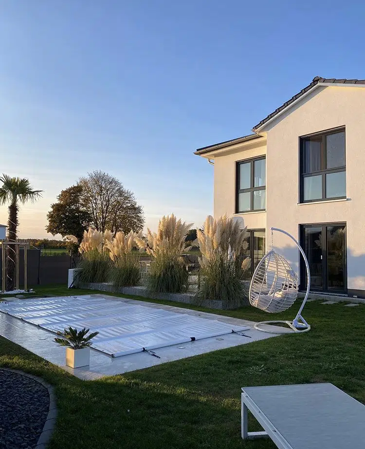 Modernes weißes Haus mit großen Fenstern, einem Garten mit Ziergräsern, einem überdachten Pool, einem weißen Hängesessel und einem Liegestuhl im Hinterhof bei Sonnenuntergang.
