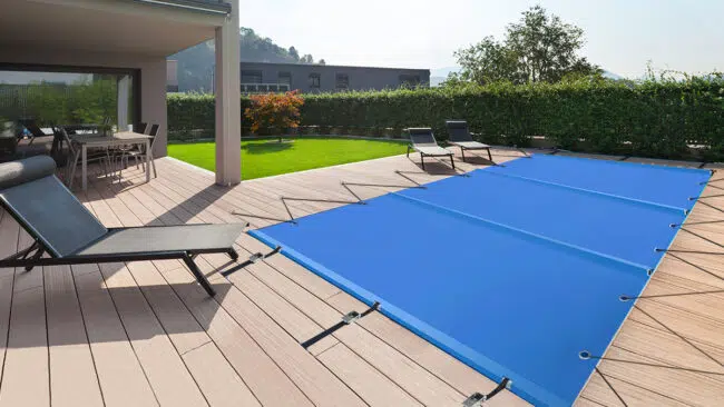 Eine Terrasse im Hinterhof mit Liegestühlen, einem Esstisch und einem rechteckigen Pool, der mit einer hellblauen Sicherheitsabdeckung abgedeckt ist, umgeben von Grünpflanzen und einer Sichtschutzhecke.