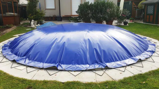 Eine große, blaue, kuppelförmige Poolabdeckung, die mit Schnüren gesichert ist, bedeckt einen runden Pool in einem Hinterhofgarten.