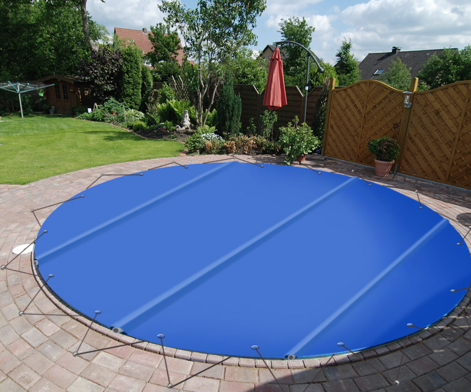Eine große blaue runde Sicherheitsabdeckung, die über einem Bodentrampolin in einem angelegten Garten mit Bäumen, Gras und einer Terrasse befestigt ist.