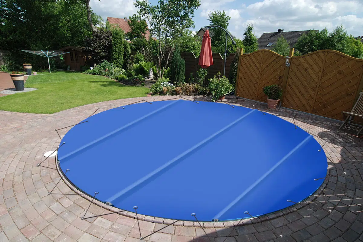 Eine große blaue runde Sicherheitsabdeckung, die über einem Bodentrampolin in einem angelegten Garten mit Bäumen, Gras und einer Terrasse befestigt ist.