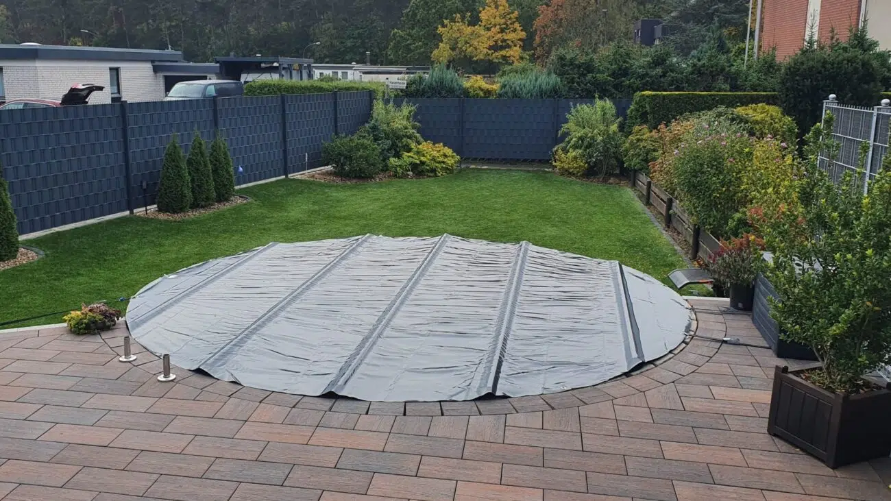 Ein runder Swimmingpool ist mit einer grauen Plane abgedeckt und liegt in einem Garten mit grünem Gras, Büschen und einer gepflasterten Terrasse.