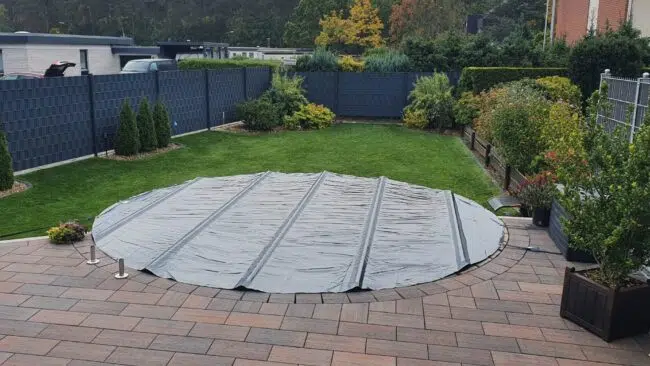 Ein runder Swimmingpool ist mit einer grauen Plane abgedeckt und liegt in einem Garten mit grünem Gras, Büschen und einer gepflasterten Terrasse.