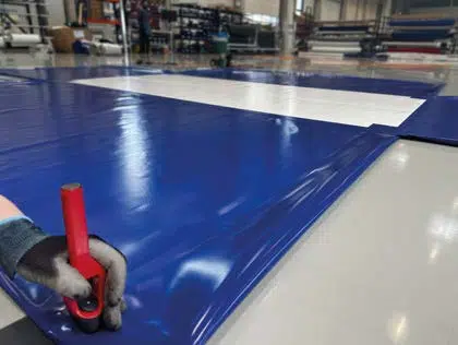 Eine behandschuhte Hand verwendet ein rotes Schneidewerkzeug, um in einem industriellen Arbeitsbereich ein großes Blatt blau-weißes Vinylmaterial zuzuschneiden.