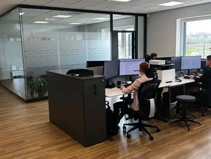 Drei Personen arbeiten an Schreibtischen mit mehreren Monitoren in einem modernen Büro mit Glaswänden, Holzboden und natürlichem Licht, das durch ein großes Fenster hereinfällt.