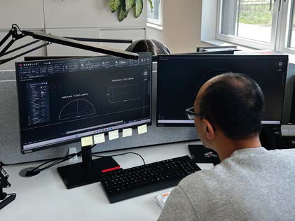 Eine Person mit Brille arbeitet an einem Schreibtisch mit zwei Computermonitoren, auf denen technische Zeichnungen und Designsoftware angezeigt werden.