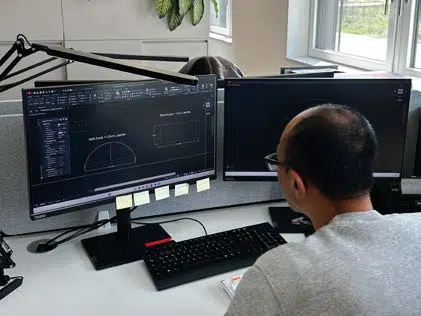 Eine Person mit Brille arbeitet an einem Schreibtisch mit zwei Computermonitoren, auf denen technische Zeichnungen und Designsoftware angezeigt werden.