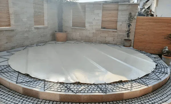 Ein runder Pool mit einer glänzenden, silbernen Abdeckung steht auf gefliesten Stufen in einem kleinen, ummauerten Hinterhof mit Topfpflanzen und einer Palme.