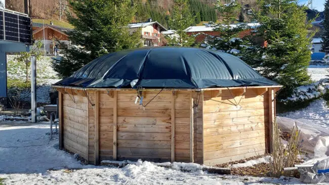 Eine runde Holzkonstruktion mit einem dunklen Planendach steht auf verschneitem Boden, umgeben von immergrünen Bäumen und nahe gelegenen Gebäuden in einer winterlichen Umgebung.