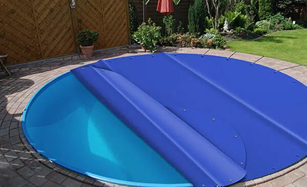 Ein runder Pool im Hinterhof ist teilweise mit einer blauen Poolabdeckung bedeckt und von einer gemauerten Terrasse, einem Holzzaun und Gartenpflanzen umgeben.