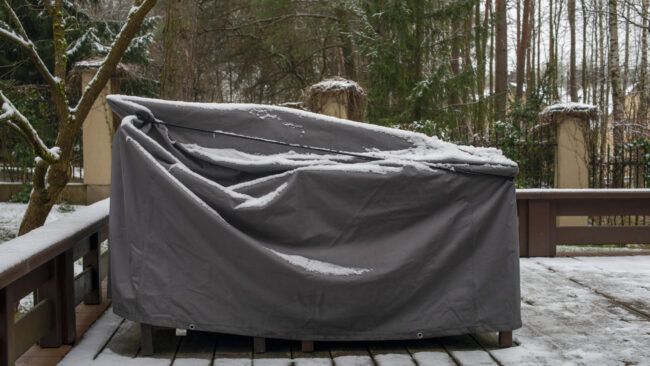 Gartenmöbel unter einer grauen Schutzhülle auf einer verschneiten Holzterrasse.