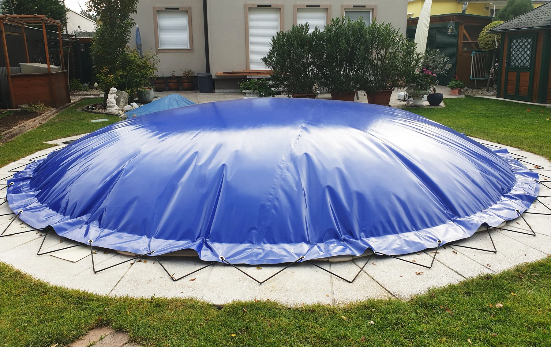Eine große, blaue, kuppelförmige Poolabdeckung, die mit Schnüren gesichert ist, bedeckt einen runden Pool in einem Hinterhofgarten.